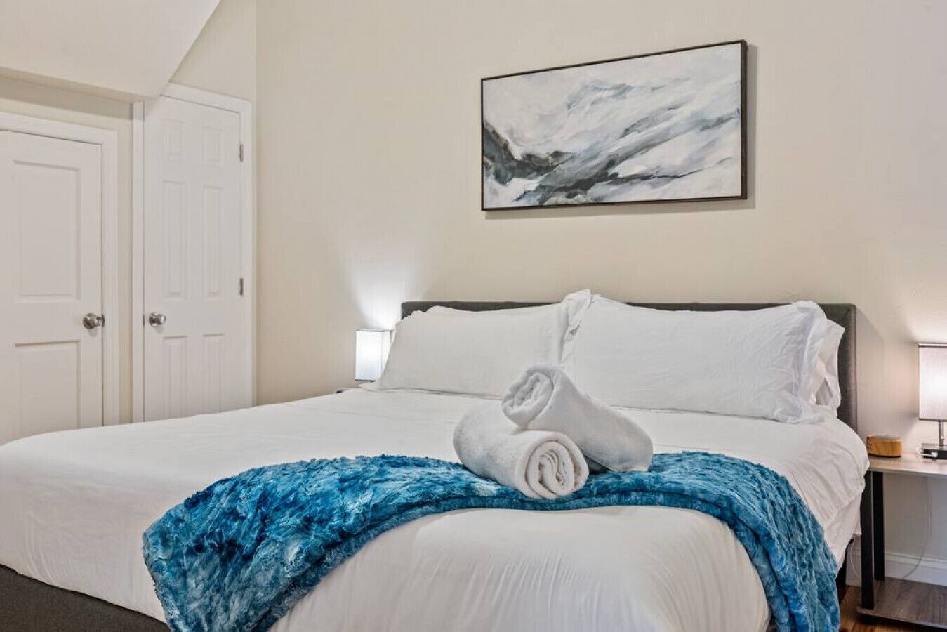Een keurig opgemaakt bed met witte lakens, een blauwe deken en opgerolde handdoeken op het bed.