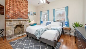 Un dormitorio con chimenea de ladrillo, cama con colcha azul y blanca, ventilador de techo y vista a la ciudad a través de ventanas amplias.