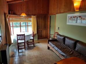 A room with a wooden floor, a brown leather sofa, a dining table with chairs, a window with curtains, and a door.