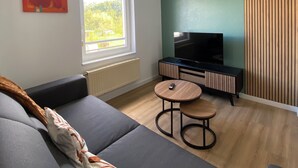 A modern living room with a grey sofa, a small wooden table, a TV, and a window with a view of greenery.