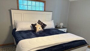 A bed with a white headboard, a blue bedspread, and a white quilt. There is a nightstand with a white lamp and a small white fan. A window with frosted glass is above the bed. - Lakeshore Cottage, For Outdoor Enthusiasts (Gaylord)