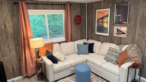 Un salon avec un canapé d’angle, une lampe, un tapis et des tableaux encadrés accrochés au mur.