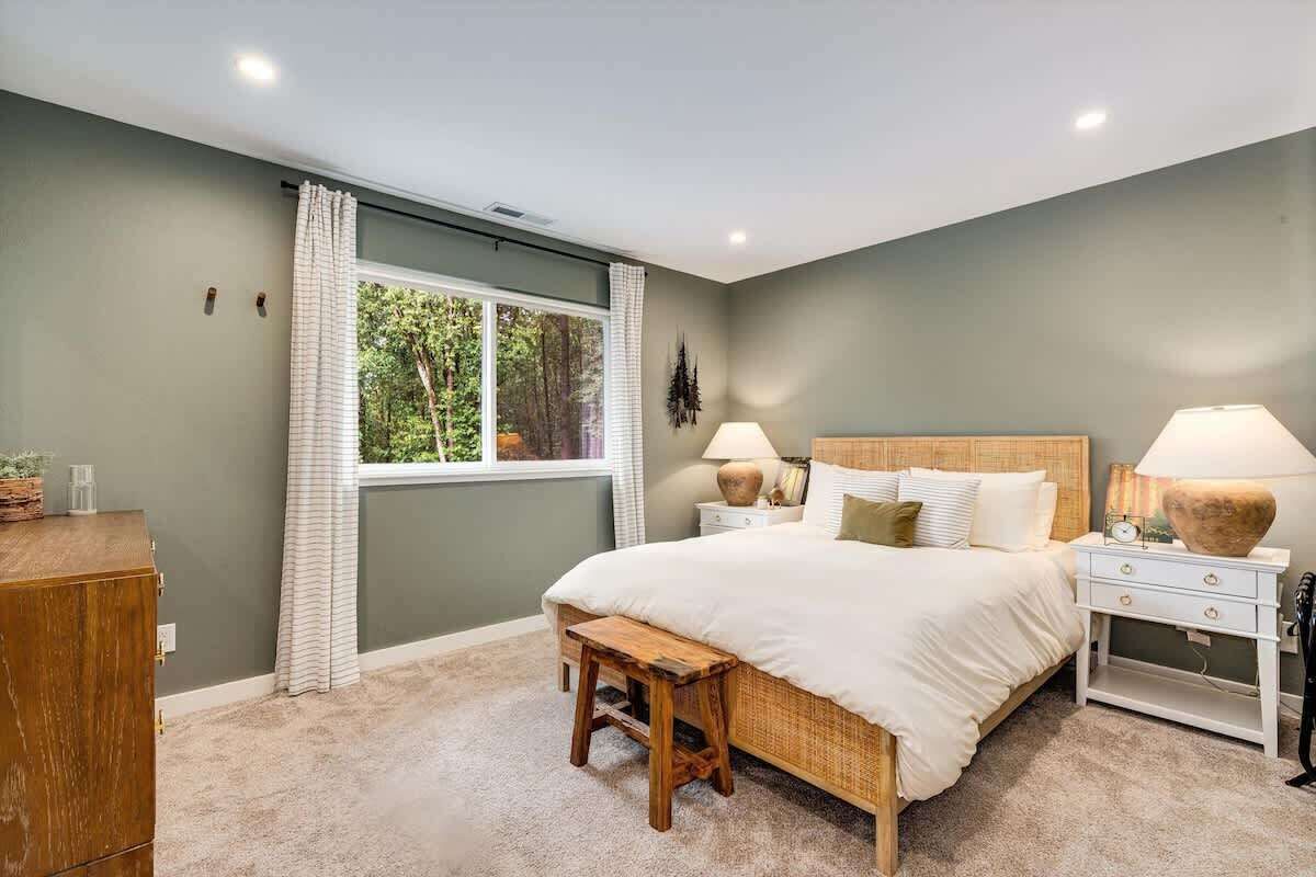 A bedroom with a large bed, two bedside tables with lamps, a wooden bench, a dresser, and a window with curtains.