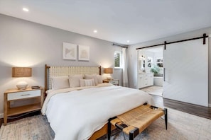 Quarto moderno com cama grande, mesas de cabeceira, um banquinho e vista para um banheiro.