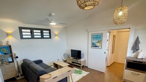 Un salon moderne avec un canapé, un téléviseur et une table basse en bois. On y trouve également un ventilateur de plafond, des lampes fixées au mur et une porte qui donne sur la cuisine.