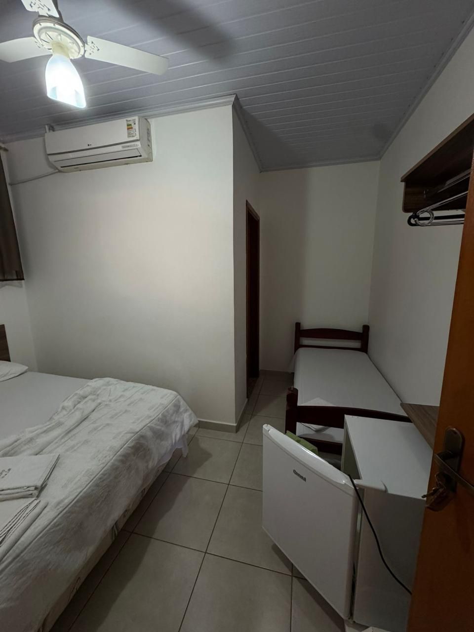 A small, compact room with a single bed, a desk, and a chair. There is an air conditioning unit on the wall and a ceiling fan.