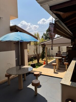 An outdoor patio area with a round table and stools, a blue umbrella, and a seating arrangement under a covered section. - Pousada Recanto Café com Leite (Olímpia)