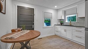 Une cuisine moderne avec des meubles blancs, une table à manger en bois et une fenêtre équipée de stores.