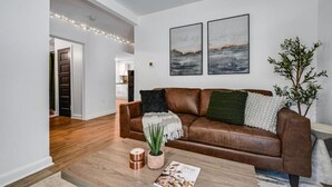 Un salon moderne avec un canapé en cuir marron, deux œuvres d’art accrochées au mur, une plante en pot et un magazine posé sur le sol.