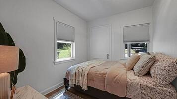 A bedroom with a bed, two windows with blinds, a lamp, and a rug.