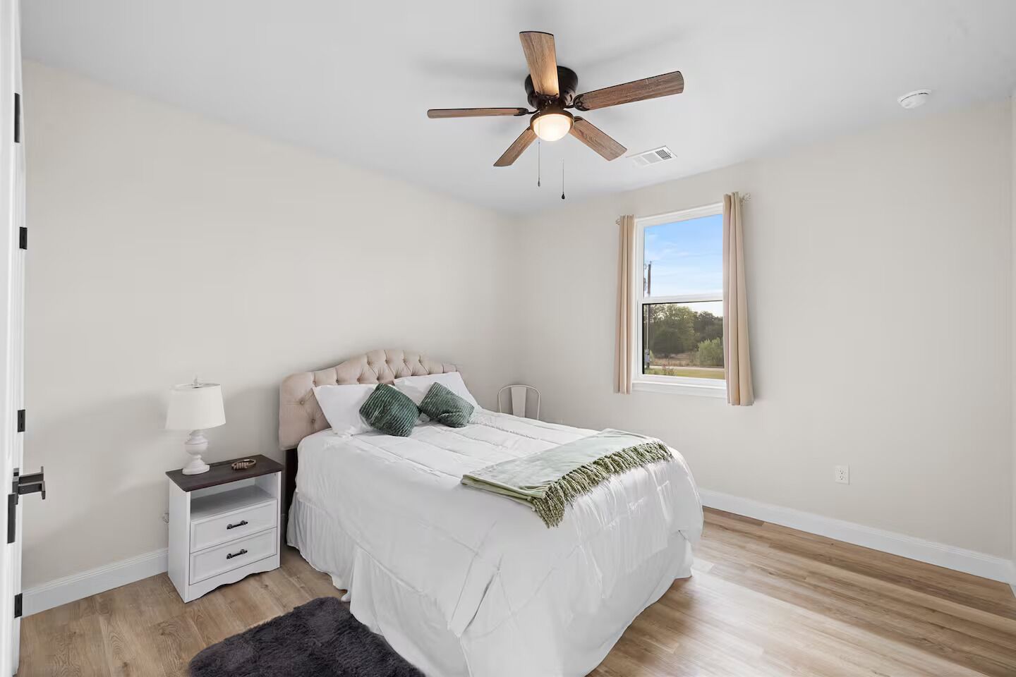 Une chambre à coucher comprenant un lit, une table de chevet, une lampe, un ventilateur de plafond, une fenêtre et une vue sur l’extérieur.