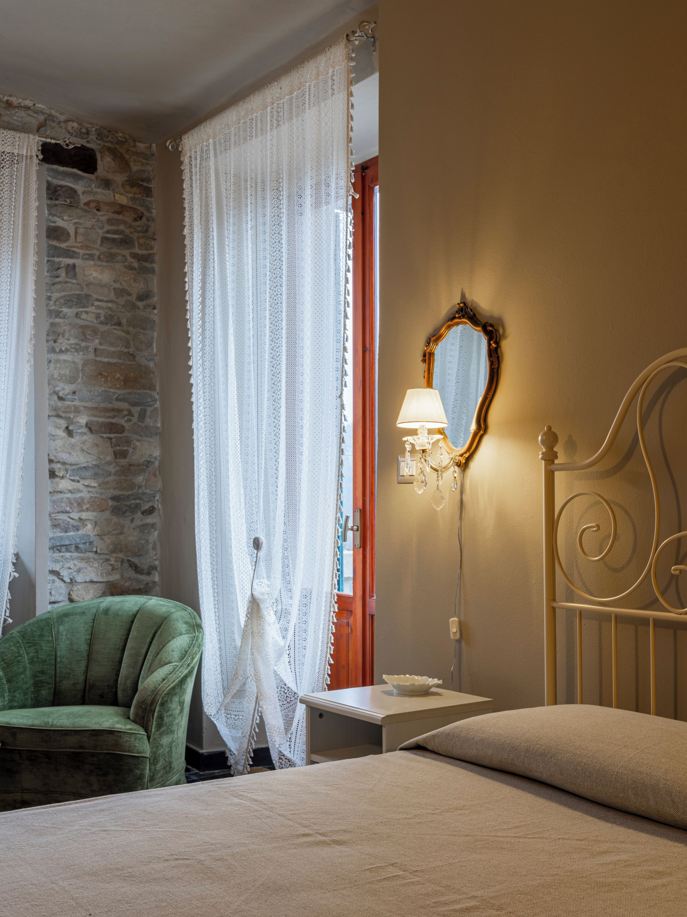 Une chambre avec un mur en pierre, un fauteuil vert, un lit avec un oreiller beige et une table de chevet avec une lampe.