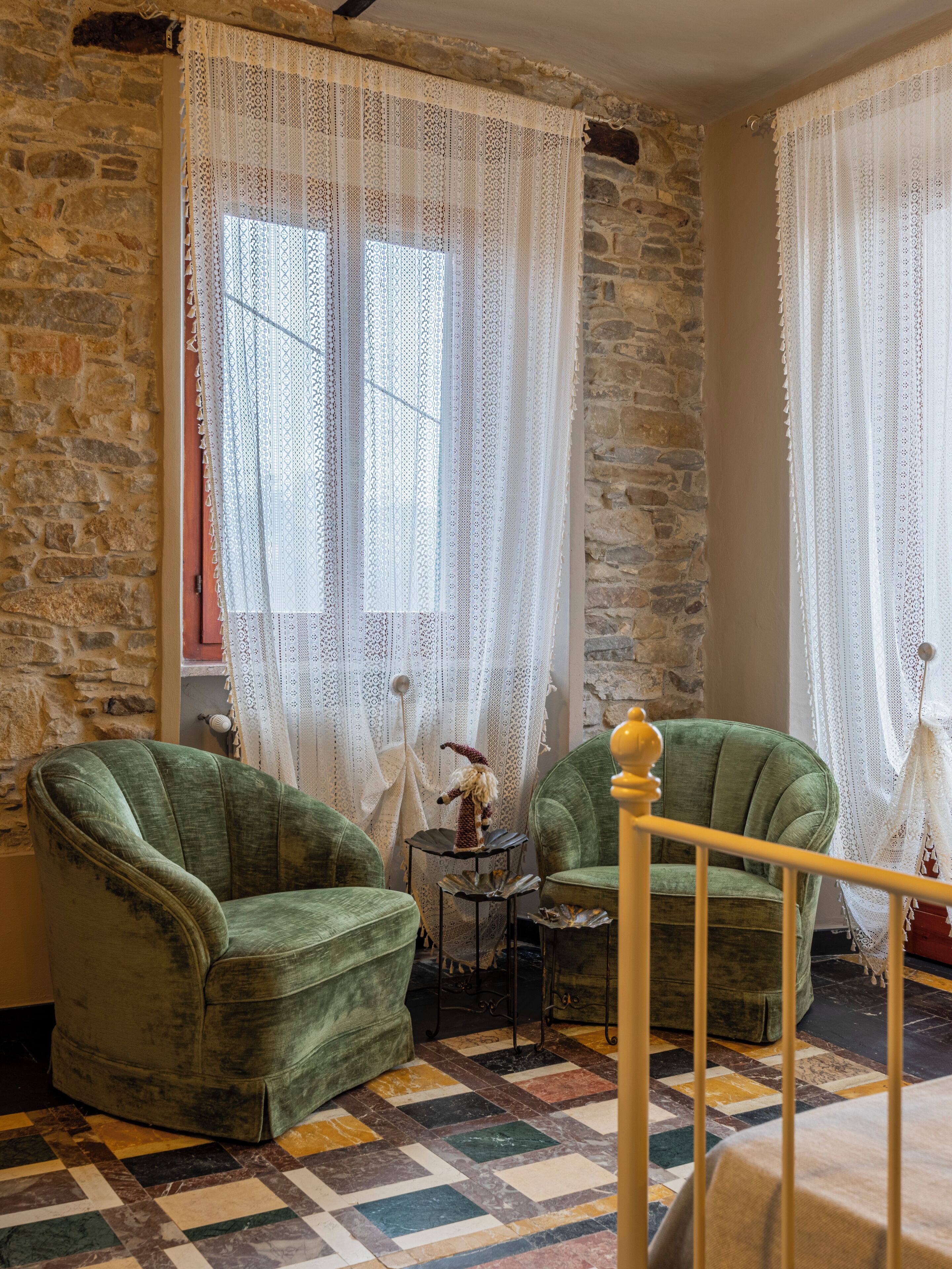 Une pièce avec un mur en pierre, deux fauteuils verts, une petite table ornée d’un objet décoratif et un lit avec une tête de lit en bois.