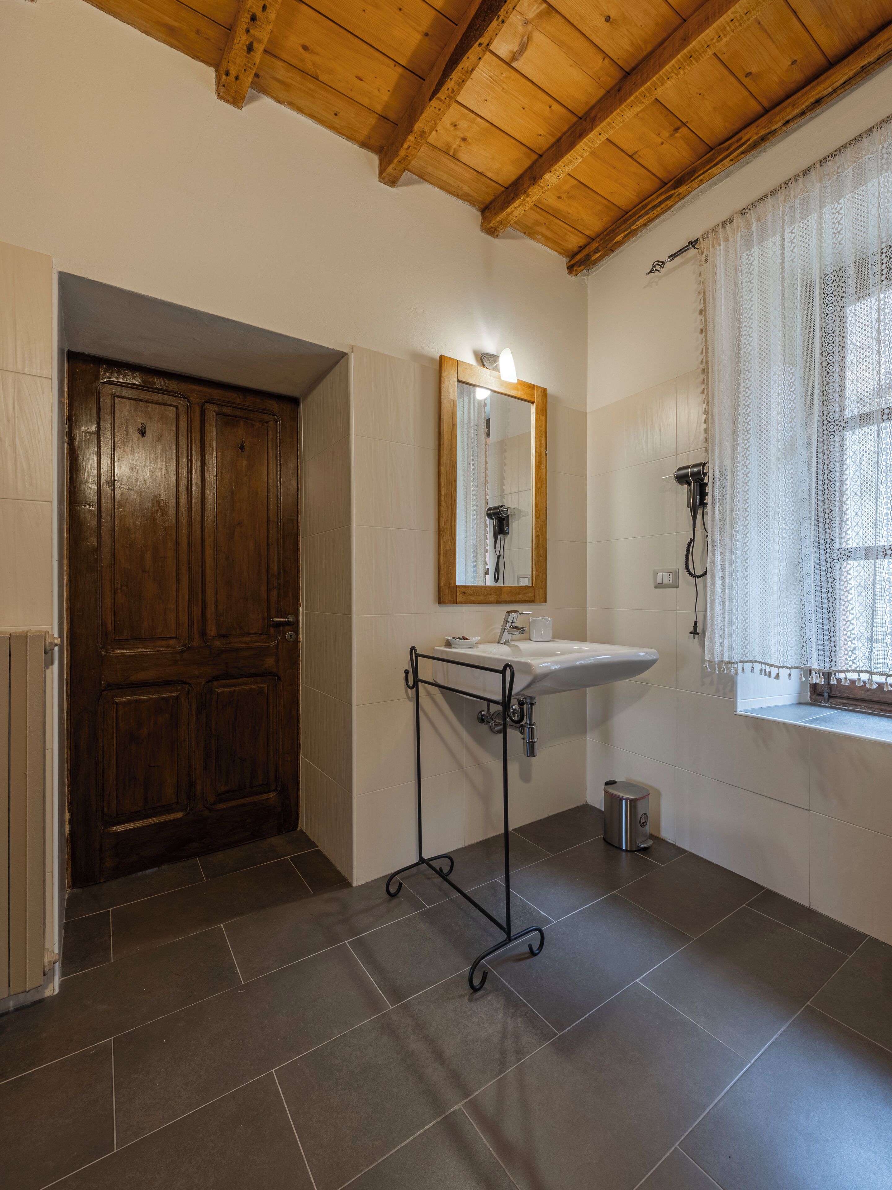 A bathroom with a wooden door, a sink with a mirror, and a window with lace curtains.