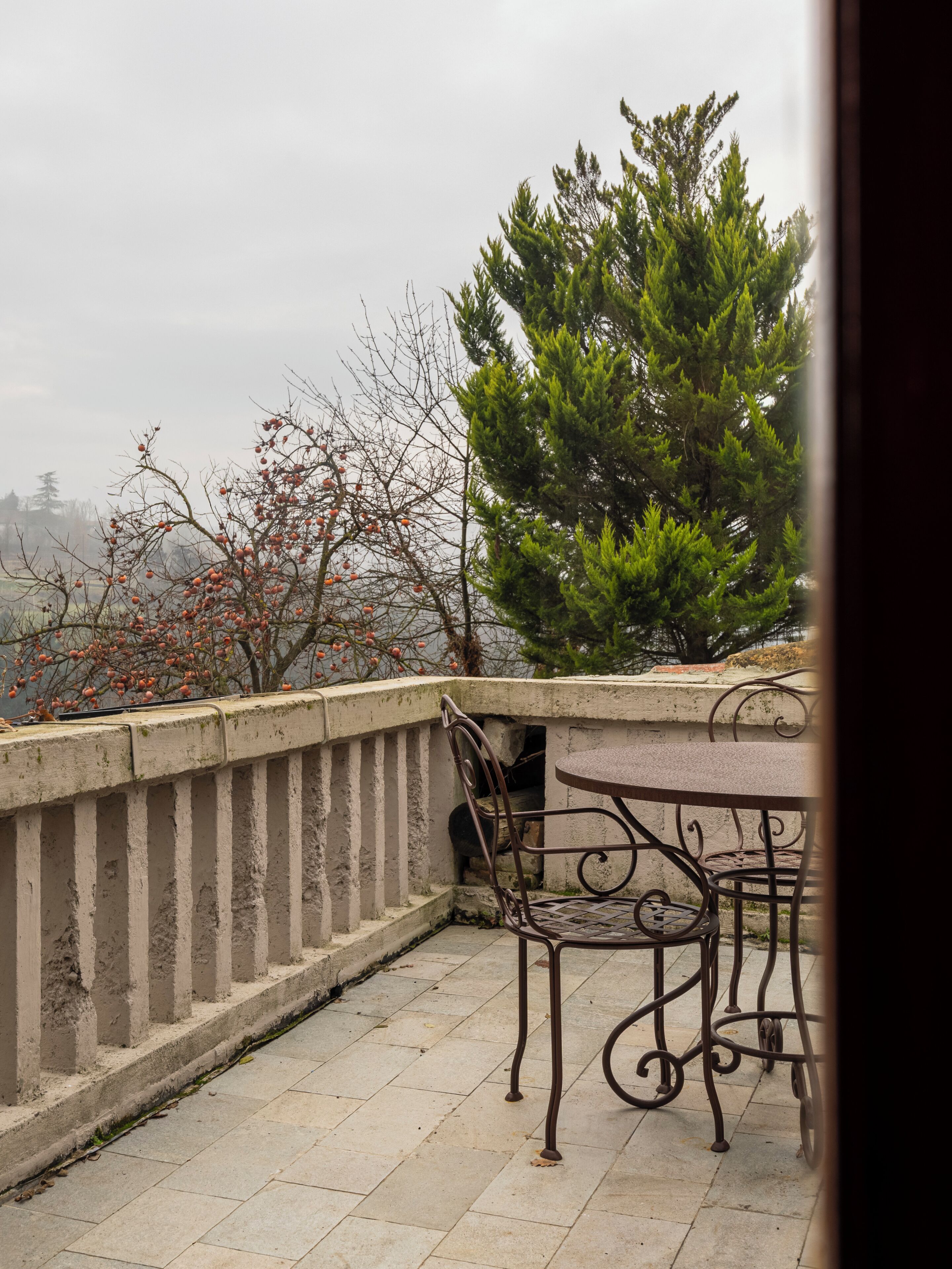 Un balcón con una mesa y sillas de metal, vista a árboles y barandilla de hormigón.