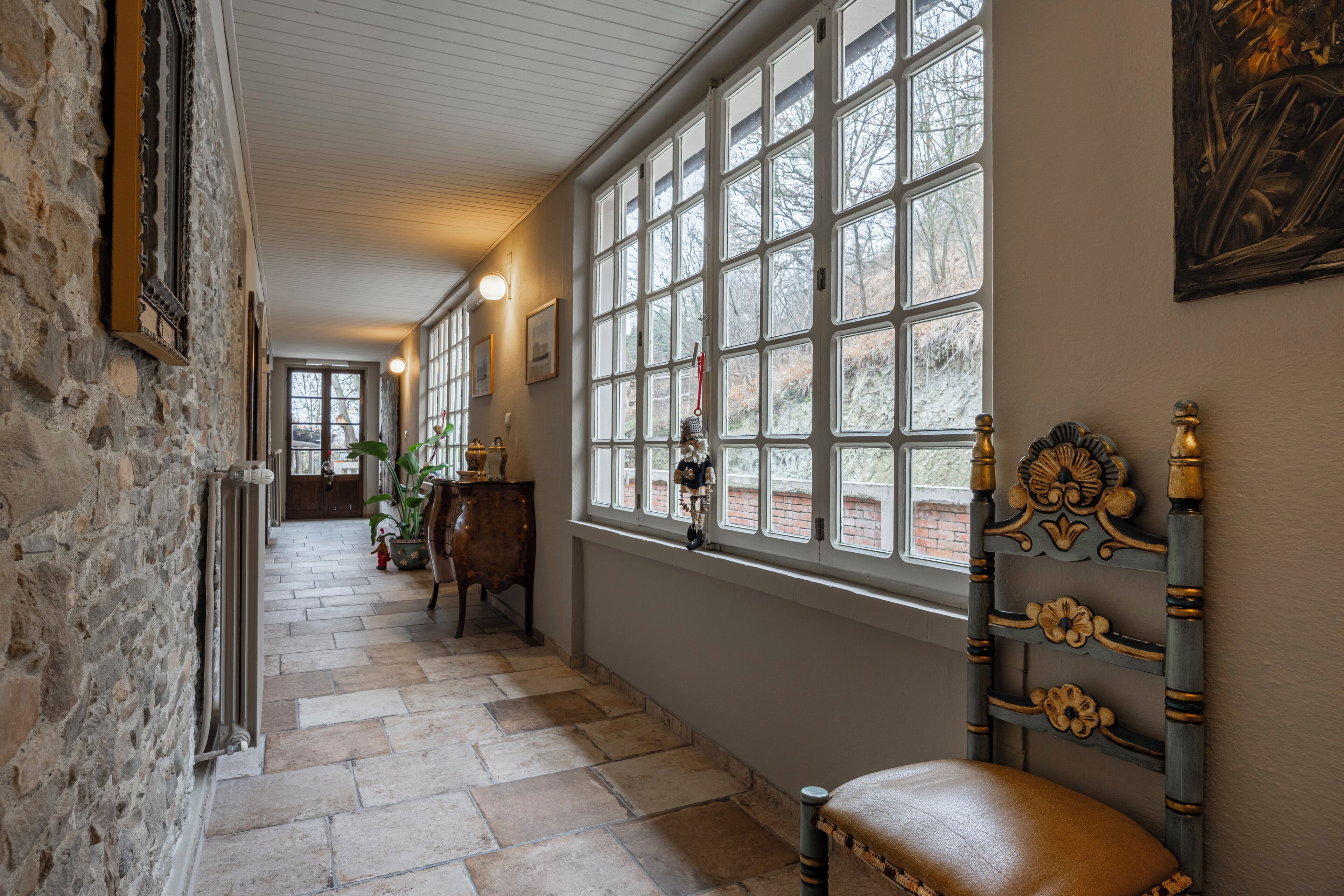 A hallway with stone walls, a wooden chair, and a large window with a view of trees.