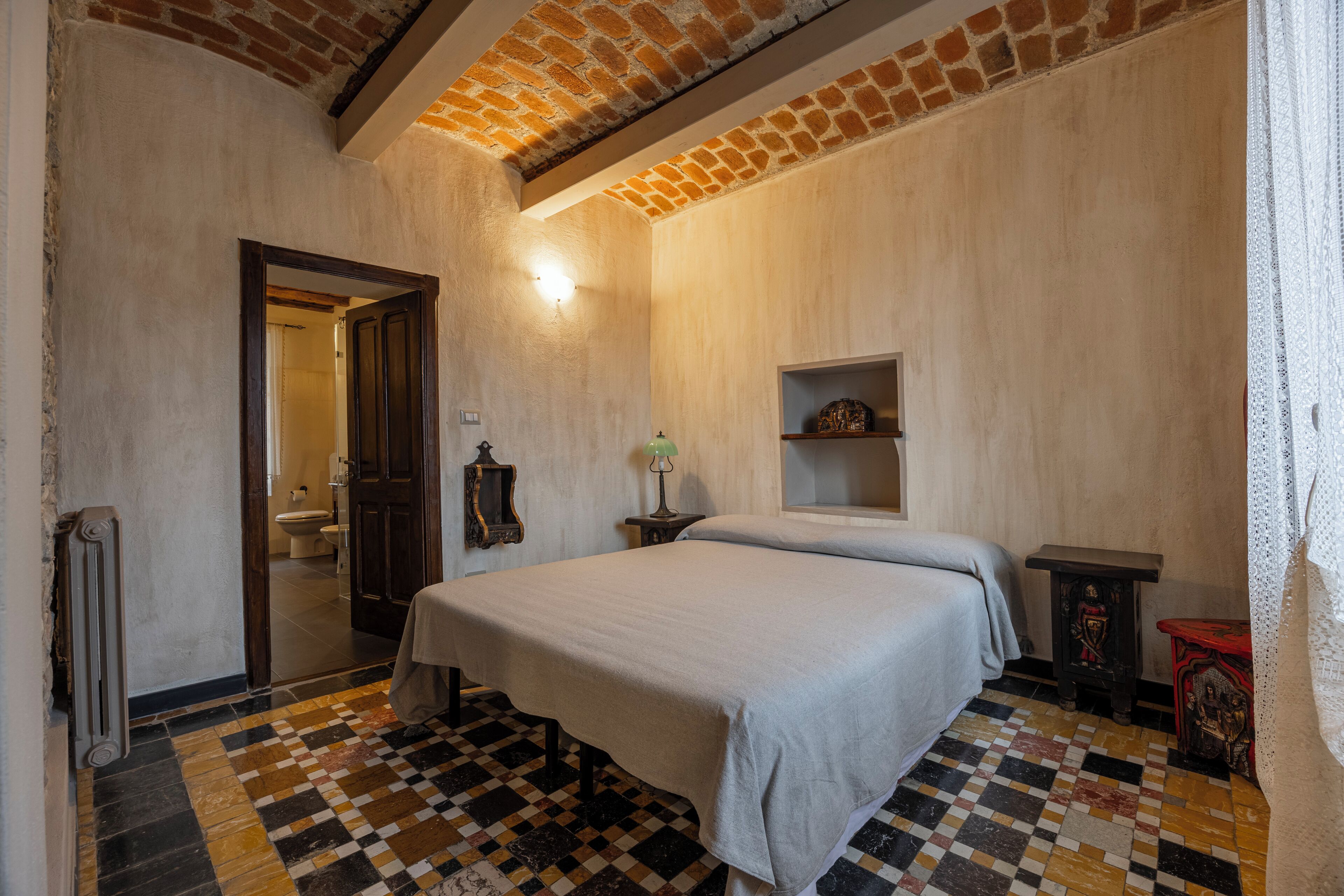 A bedroom with a checkered floor, a bed, a radiator, a wooden door, and a shelf with a lamp.