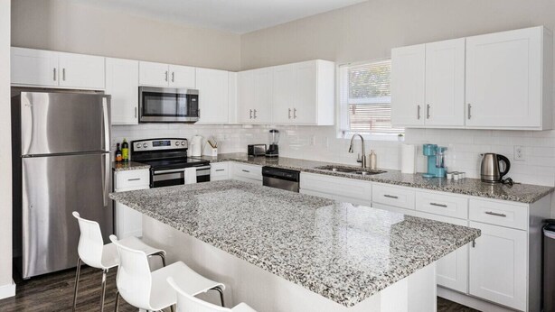 Una cocina moderna con armarios blancos, electrodomésticos de acero inoxidable, encimeras de granito y una barra de desayuno con sillas blancas.