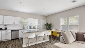 Una cocina moderna con armarios blancos, encimera de granito y electrodomésticos de acero inoxidable. Hay una zona de comedor con una mesa amarilla y sillas blancas.