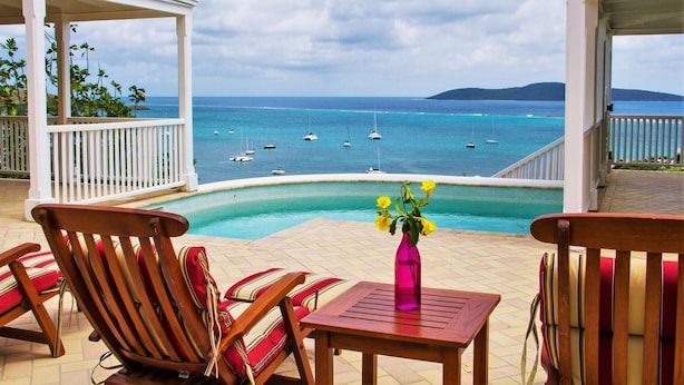 A poolside area with wooden chairs and a table, overlooking a clear blue sea with boats. - A Yacht Haven (Christiansted)