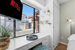 A modern bedroom with a large window, a bed, a desk with a chair, and a small shelf with decorative items.