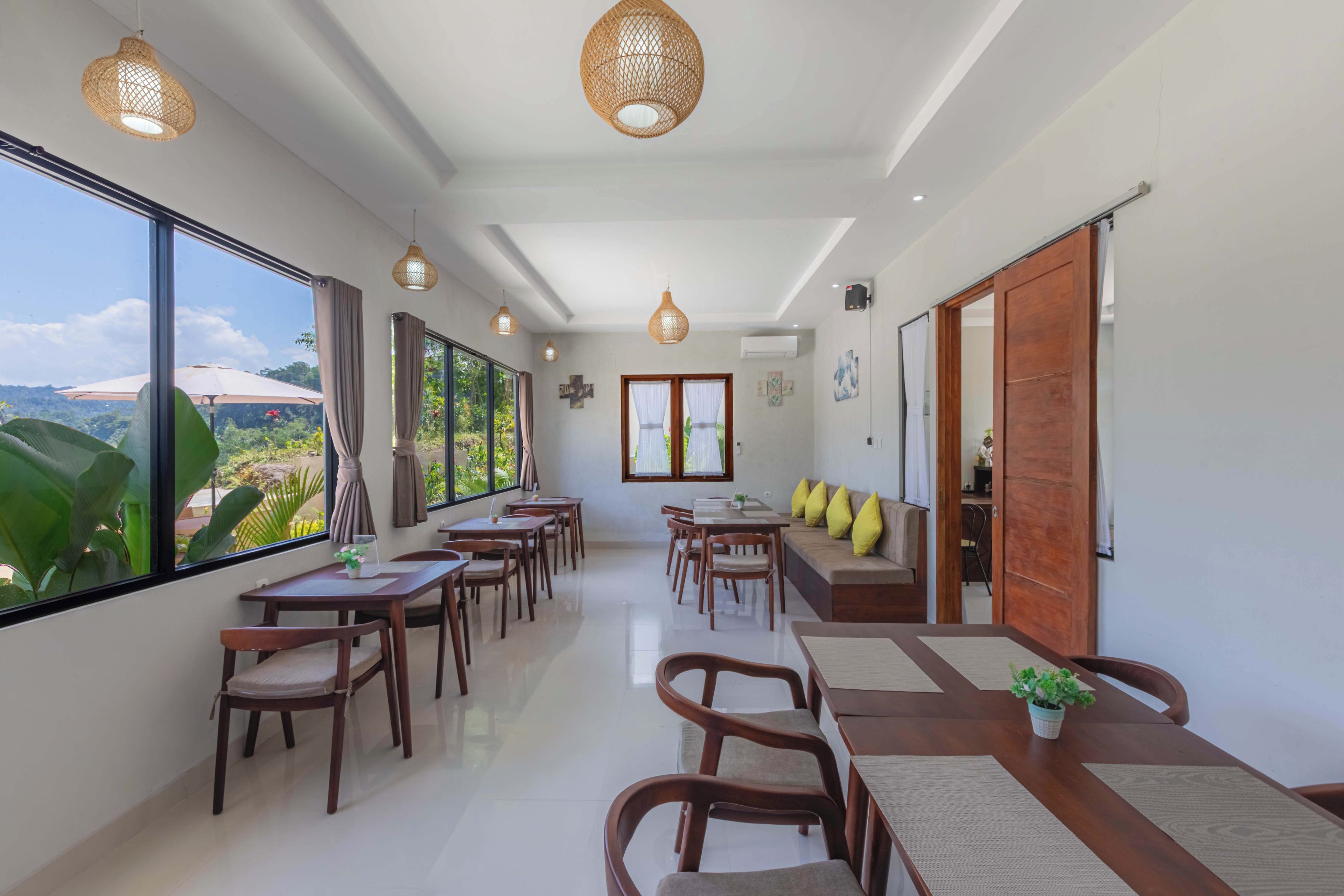 A spacious dining area with wooden tables and chairs, large windows offering a view of greenery, and a ceiling with woven light fixtures.