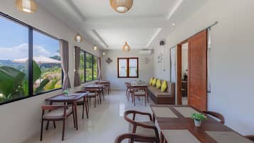 A spacious dining area with wooden tables and chairs, large windows offering a view of greenery, and a ceiling with woven light fixtures.
