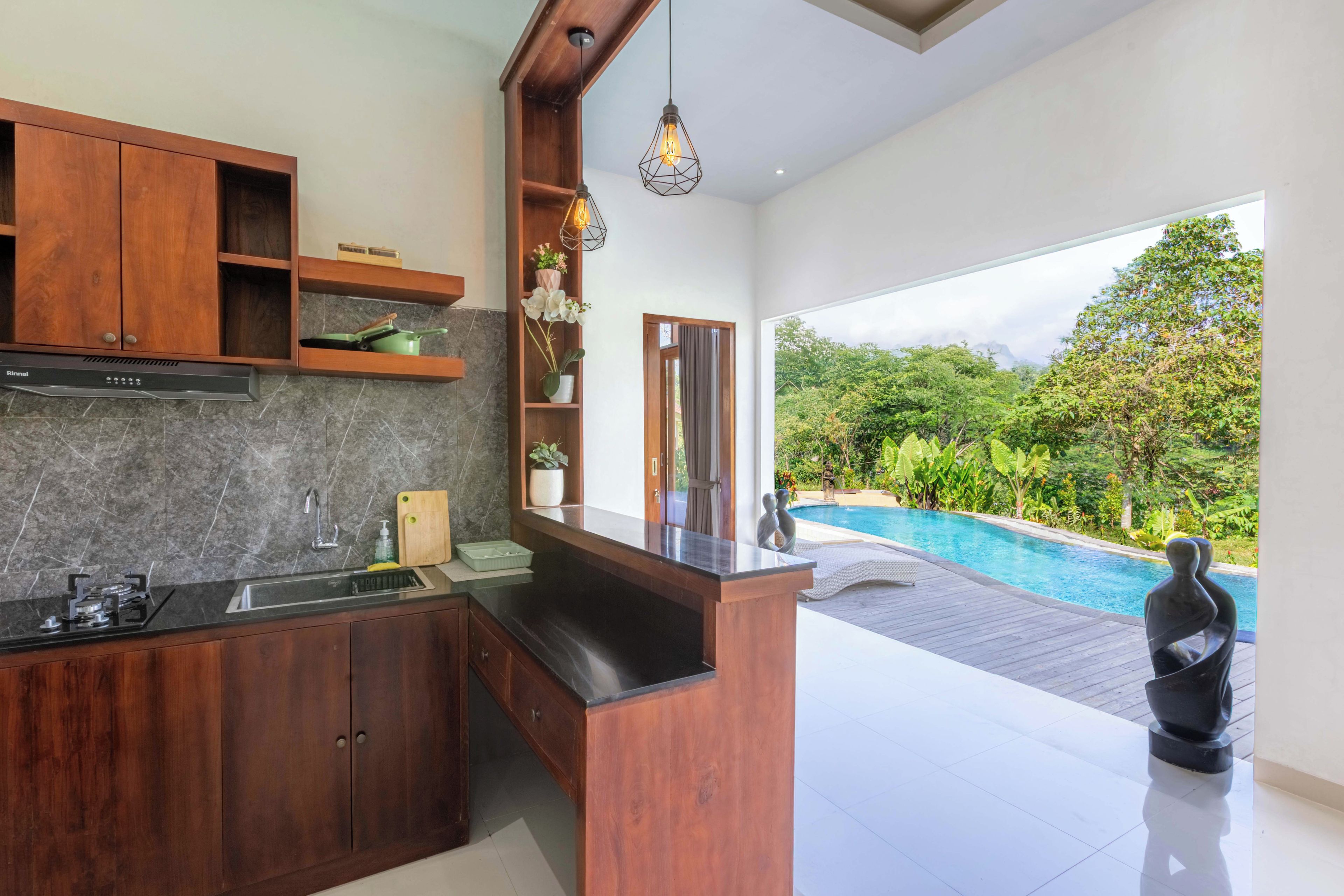 A modern kitchen with a built-in stove, a sink, and wooden cabinets. There is a view of a swimming pool and lush greenery outside.