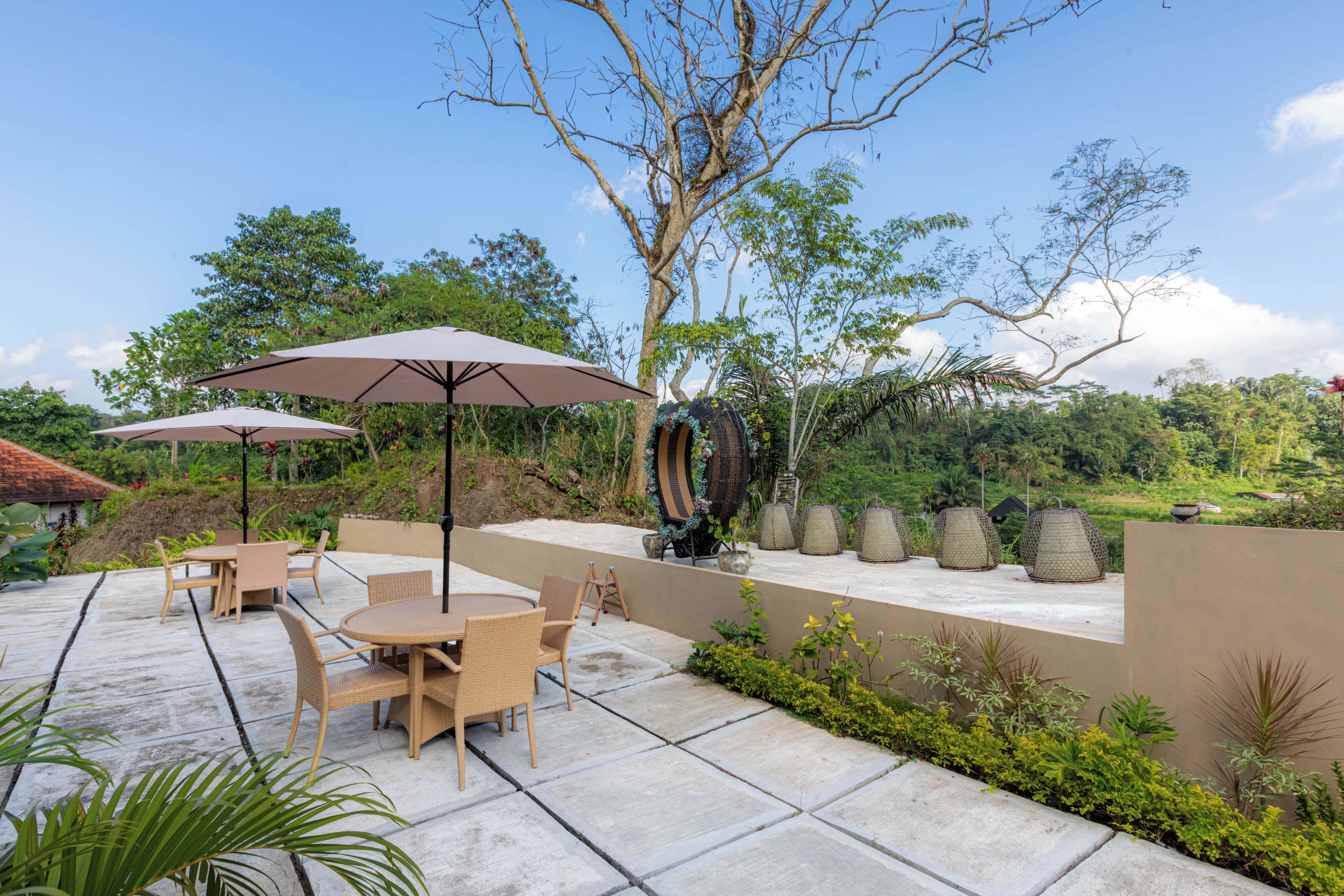 Um pátio externo com mesas e cadeiras, cercado por vegetação e céu claro.