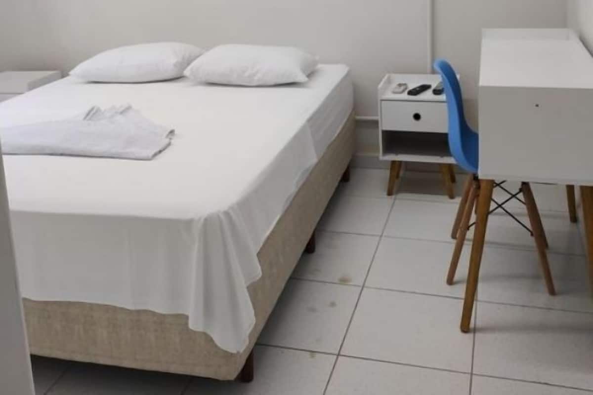 Una cama individual con ropa de cama blanca, una mesita de noche pequeña con un teléfono y cargador, y un escritorio con una silla.