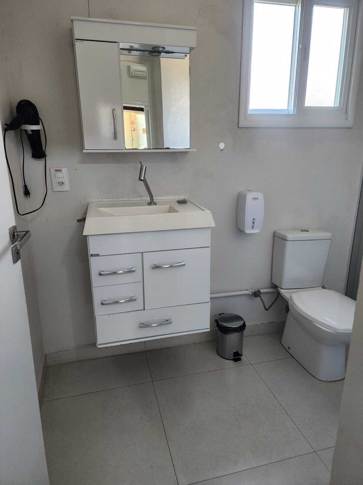 Casa de banho com uma pia branca, espelho e sanita. Há um cesto de lixo e um secador de mãos na parede.