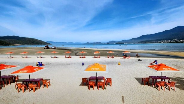 Ein Strand mit Reihen von orangefarbenen Sonnenschirmen und Plastikstühlen, einem klaren Himmel und fernen Bergen.