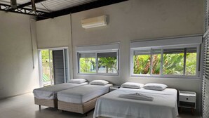 Ein Zimmer mit drei Betten, großen Fenstern mit Jalousien, einer Klimaanlage und einem Fliesenboden.