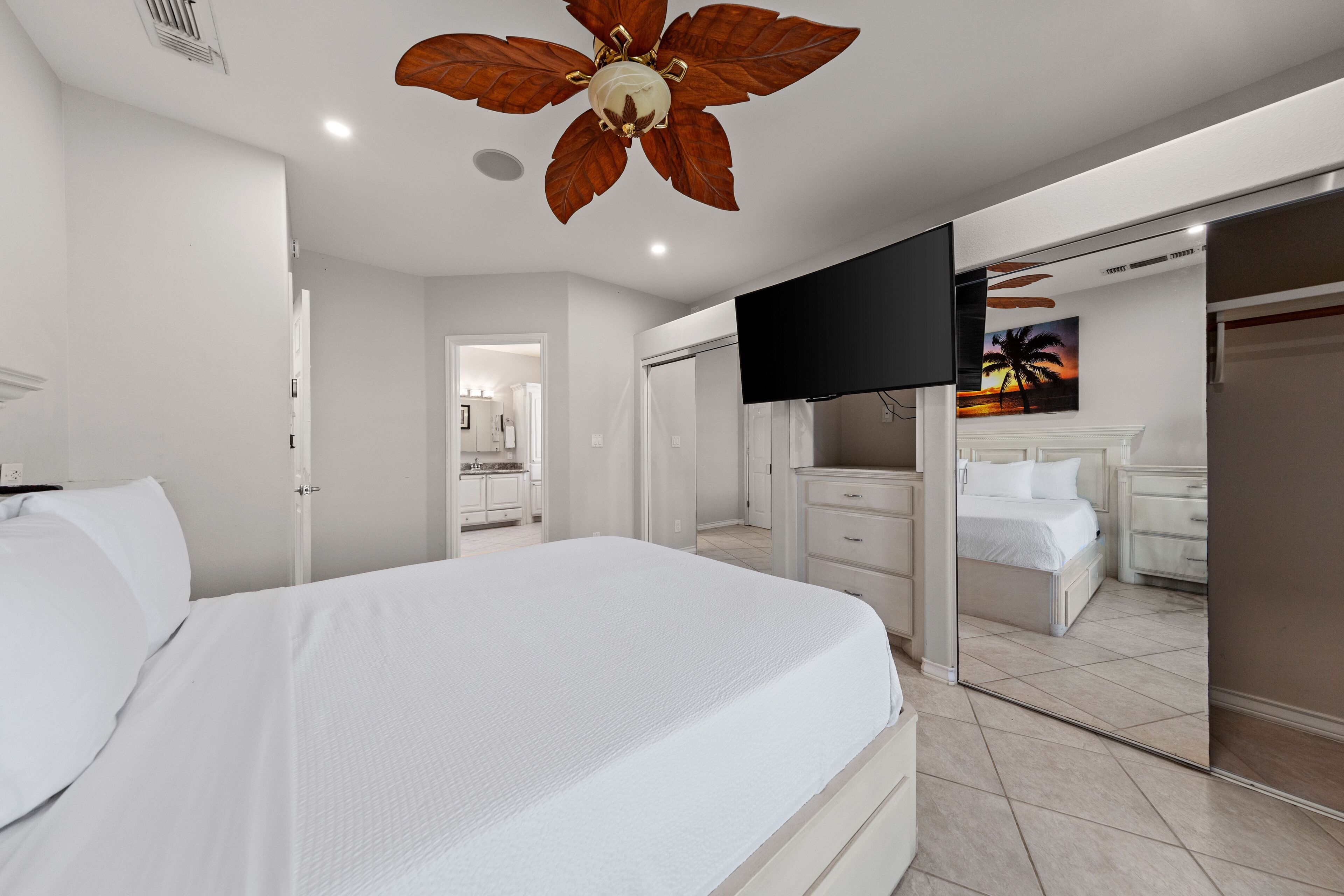 Una camera da letto moderna con un letto grande, una ventola a soffitto, un televisore e un bagno visibile attraverso una porta aperta.