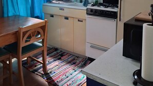 Uma cozinha compacta com armários vermelhos, uma geladeira branca, um fogão e um micro-ondas. Há uma mesa de madeira com cadeiras e um tapete colorido no chão.