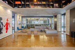 A modern lobby with a wooden floor, a grey sectional sofa, and a glass wall. - Your Complete Stay in a Building With a Pool Just Steps From the Subway - FRE (São Paulo)