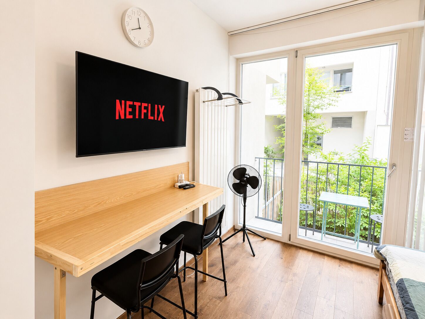 A modern room with a wooden desk, two black chairs, a TV mounted on the wall displaying 'NETFLIX', a standing fan, and a sliding glass door leading to a balcony with greenery.