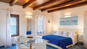 A bedroom with a bed, a glass table, white chairs, a painting, and a view of the sea.
