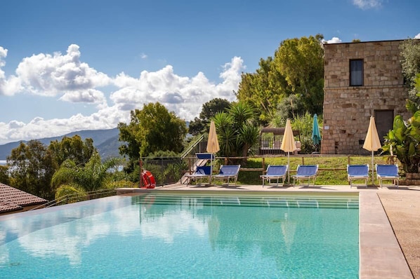 A swimming pool with lounge chairs and umbrellas, surrounded by greenery and a stone building.