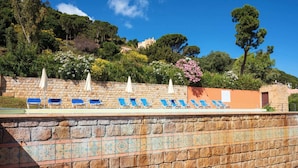 A stone wall with blue lounge chairs and white umbrellas, overlooking a hillside with trees and buildings.
