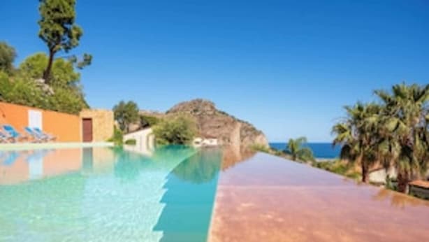 A long, narrow swimming pool with clear water, surrounded by palm trees and a rocky hill in the background.