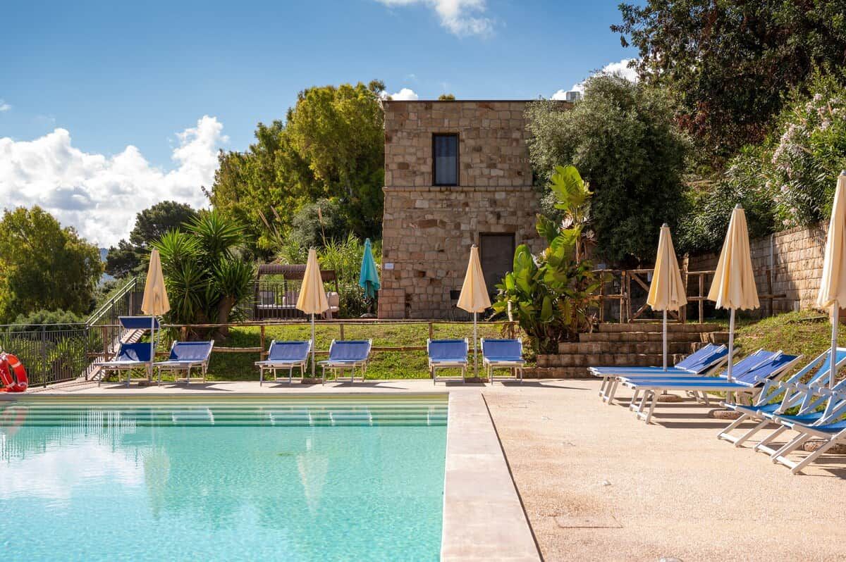 Una piscina con lettini e ombrelloni, immersa nel verde e affiancata da un edificio in pietra.