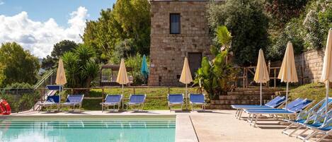 Ein Swimmingpool mit Liegestühlen und Sonnenschirmen, umgeben von Grünflächen und einem Steinhaus.