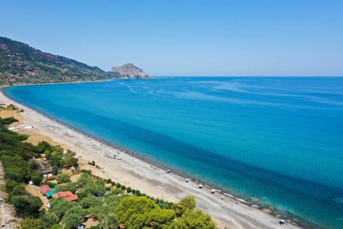 A coastal landscape with a beach, clear blue sea, and a distant hill.