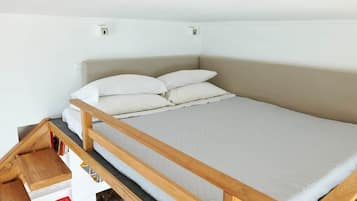 Un lit simple avec un cadre en bois et une literie blanche, dans une pièce aux murs blancs et agrémentée d’une petite étagère avec des livres.