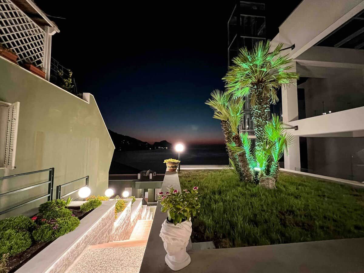 Un espacio exterior nocturno con una escalera que lleva a un balcón, una palmera y vista al mar.