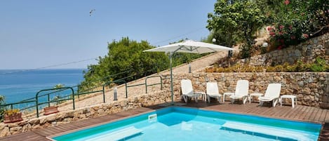 Une piscine avec des chaises longues et un mur en pierre, donnant sur une étendue d’eau.