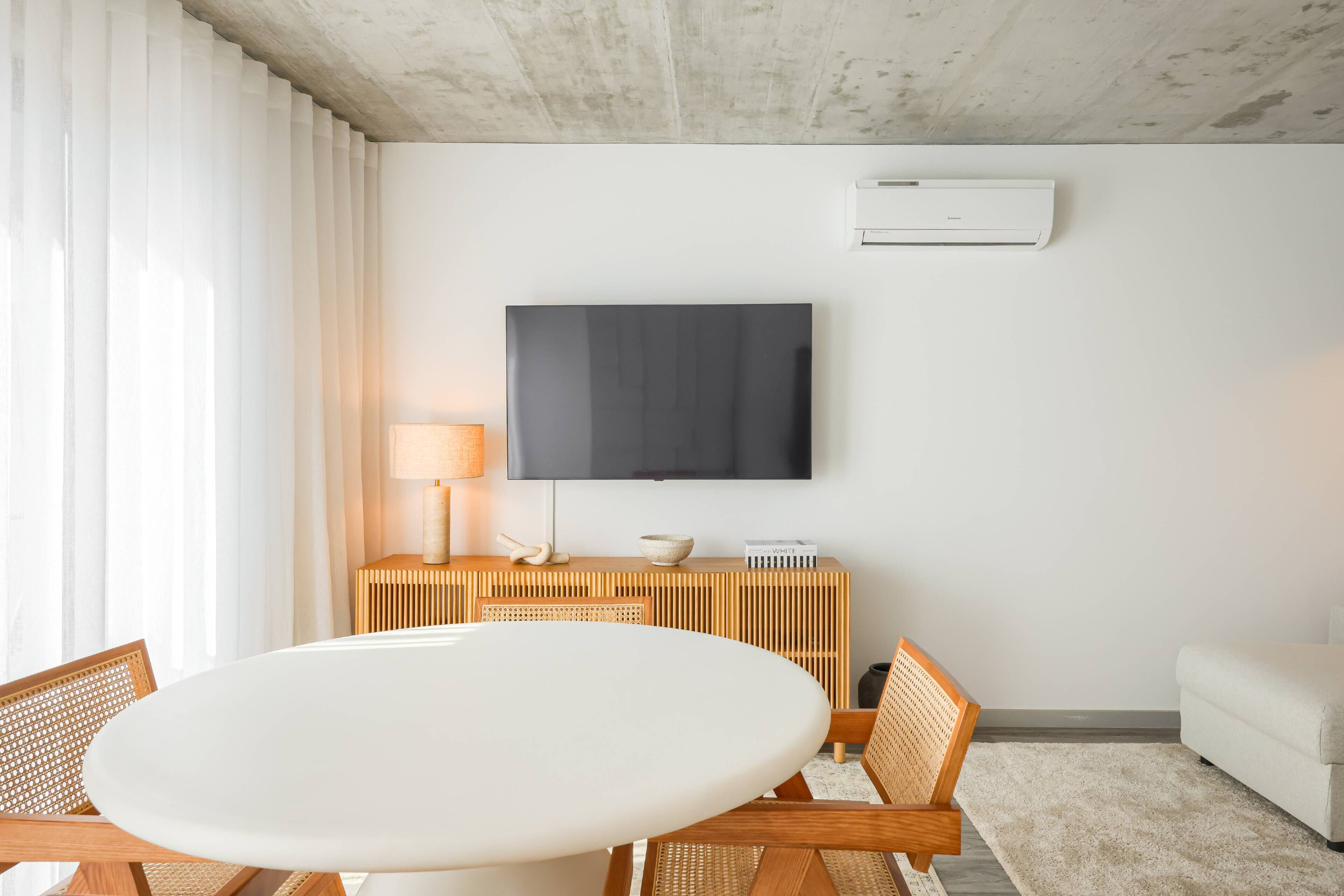 A modern living room with a round table, wooden chairs, a flat-screen TV mounted on the wall, and a white sofa.