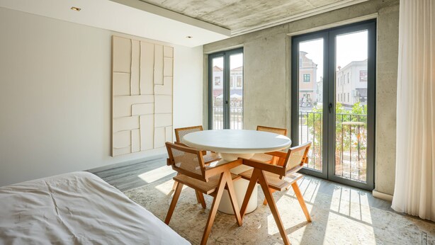 Quarto de hotel moderno com uma cama grande, uma mesa de jantar redonda com cadeiras, uma janela ampla com vista para edifícios e um teto de concreto.