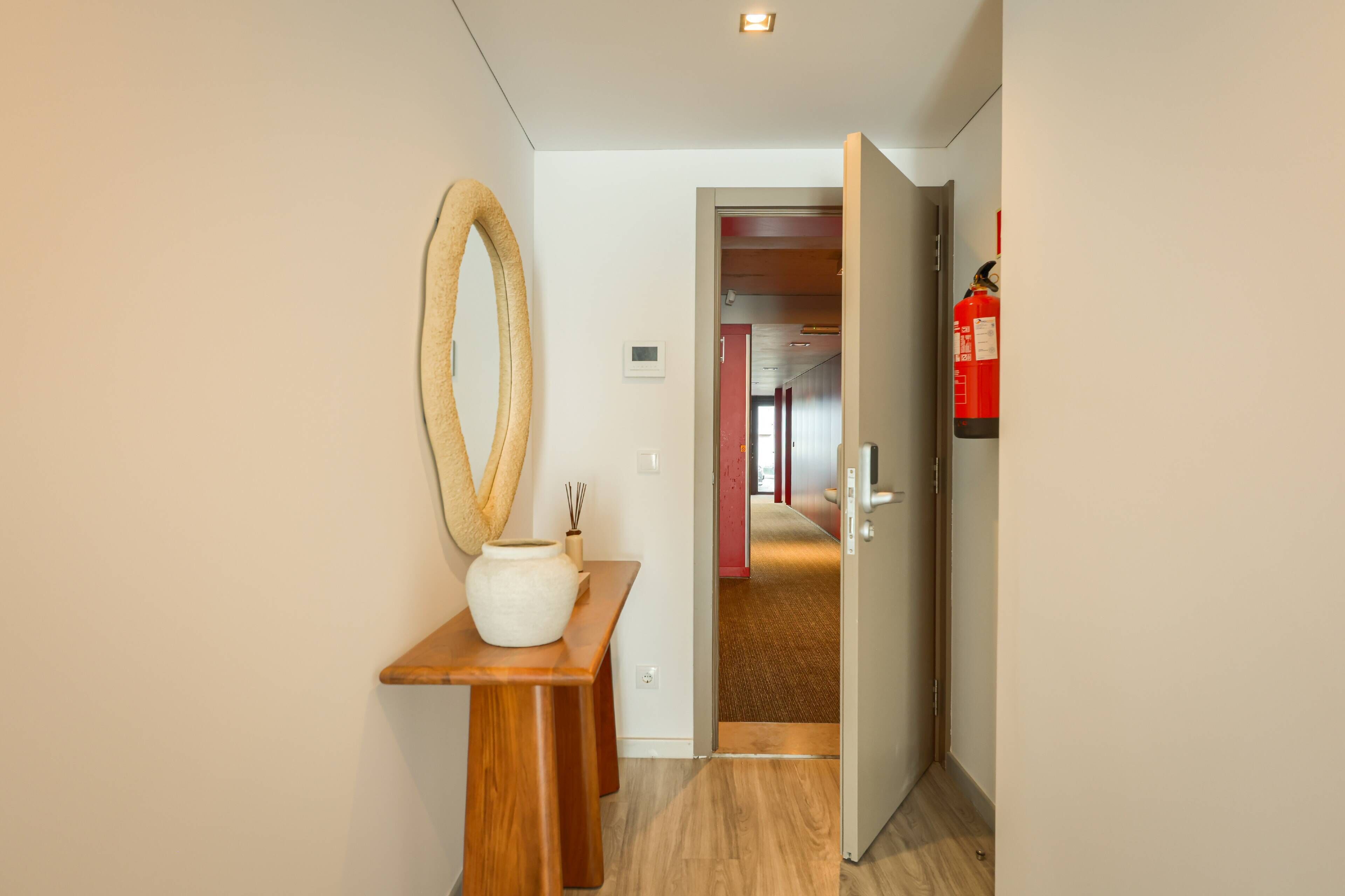 A hallway with a wooden bench, a large mirror, and a vase. A fire extinguisher is mounted on the wall.